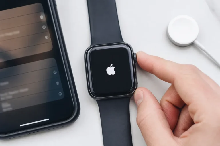 Apple Watch تعرض شعار Apple الأبيض مع إصبعين قرب Digital Crown والزر الجانبي وهاتف وتوصيلة شحن بجانبها